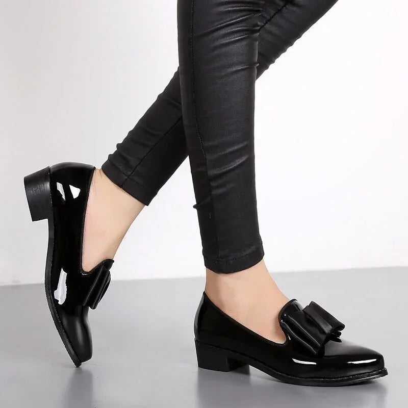 A person wearing shiny black patent leather loafers with a statement bow and low block heel, paired with black fitted pants, stands on tiptoe against a light gray background.