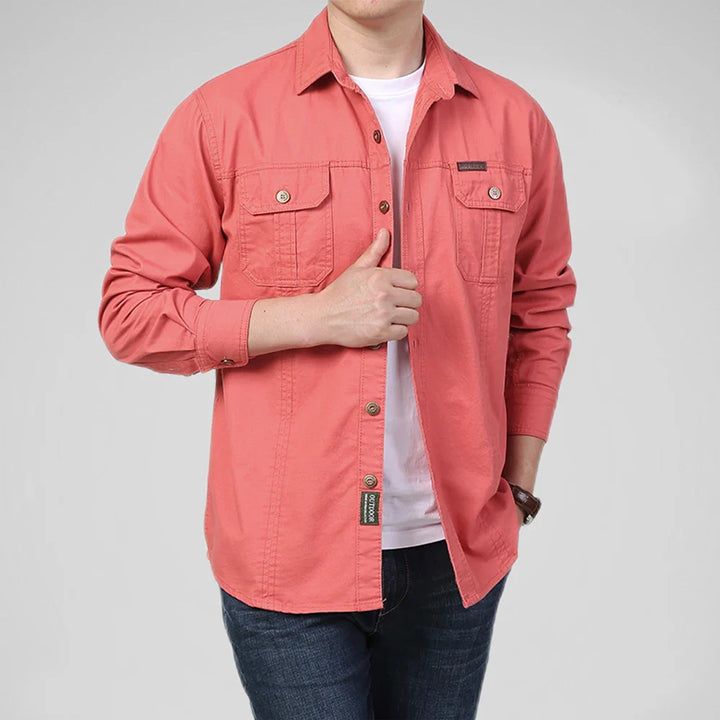 A person wearing a relaxed fit, coral pink 100% cotton shirt over a breathable undershirt and dark jeans, with one hand touching the shirts buttons and the other in their pocket, stands against a plain light background.