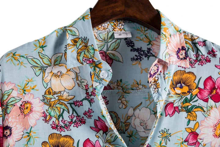 A close-up of a men's genuine linen shirt in light blue, featuring a vibrant floral pattern in pink, yellow, orange, and green, hanging on a wooden hanger for breathable comfort.
