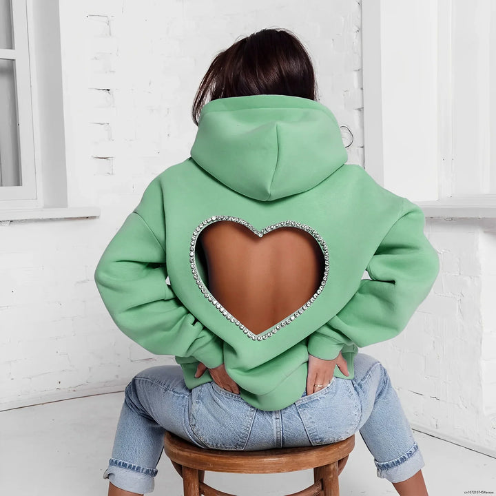 A person wearing an oversized mint green heart-shaped open back hoodie with a rhinestone trim sits on a stool facing a white brick wall. Light blue jeans complete the look.