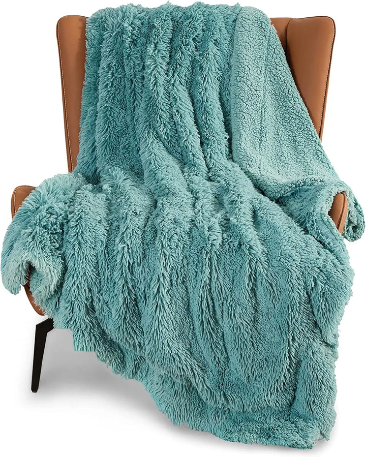 A soft, sage green faux fur throw blanket draped over a brown leather armchair with angled arms and black legs.