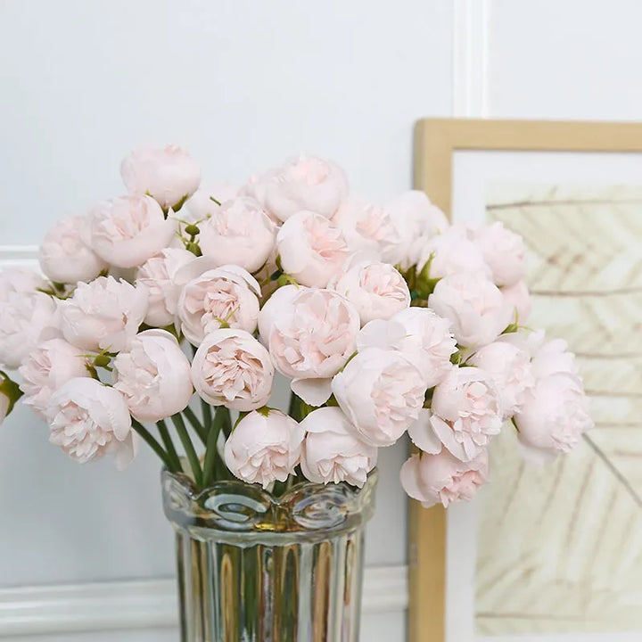 A glass vase filled with light pink peony flowers, featuring realistic faux petals, is placed in front of a framed artwork with neutral tones, against a white paneled wall.