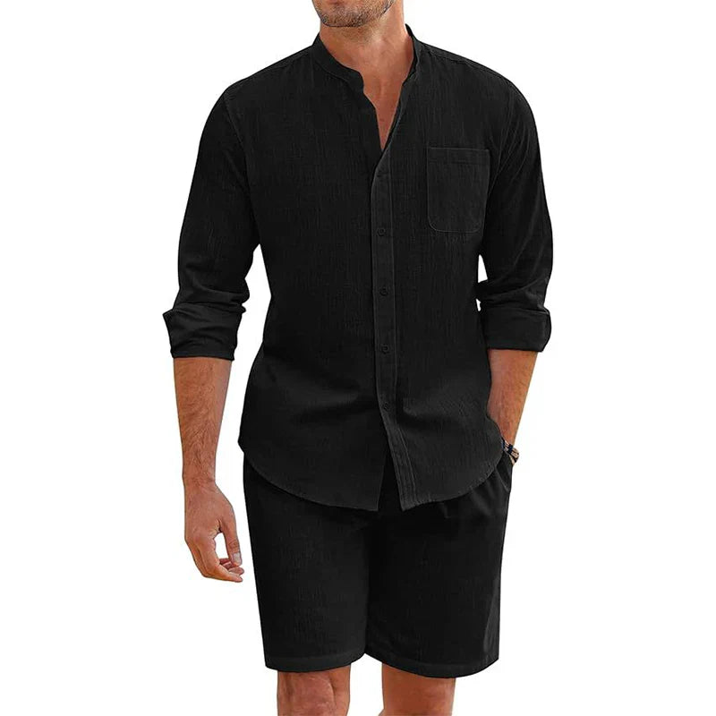 A man wearing a black, vintage stand collar shirt with sleeves rolled up and matching baggy shorts. He has one hand in his pocket; the cotton linen outfit features a single chest pocket on the shirt.