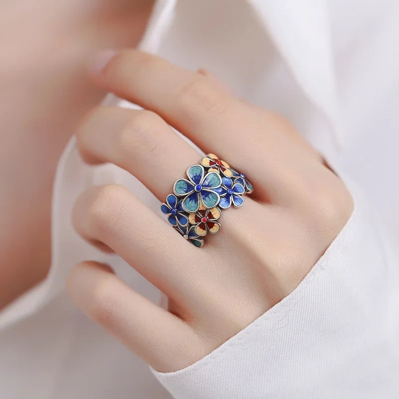 A hand wearing a white shirt displays a colorful ring, crafted with vibrant colors and blue and orange enamel flowers, reminiscent of stained glass.
