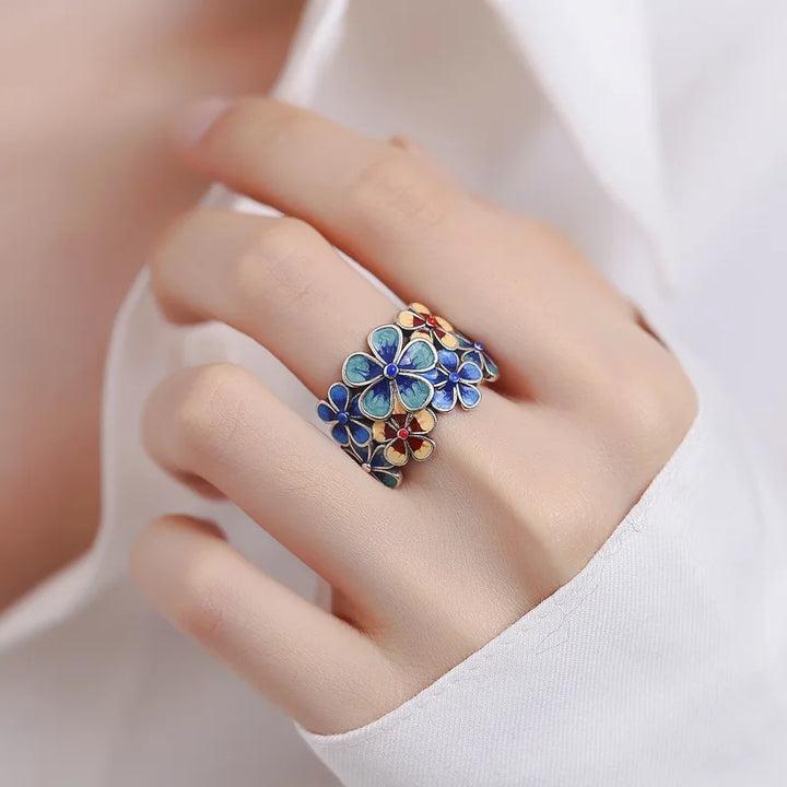 A hand wearing a white shirt displays a colorful ring, crafted with vibrant colors and blue and orange enamel flowers, reminiscent of stained glass.