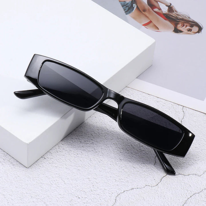 Black retro rectangle sunglasses with a lightweight frame and dark UV400 protection lenses, placed on a textured light surface beside glossy white and photographic prints of a woman in a red bikini.