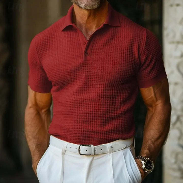 A man wearing a classic polo in textured burgundy knit fabric, tucked into high-waisted white trousers with a white belt and a watch on his left wrist, standing outdoors.