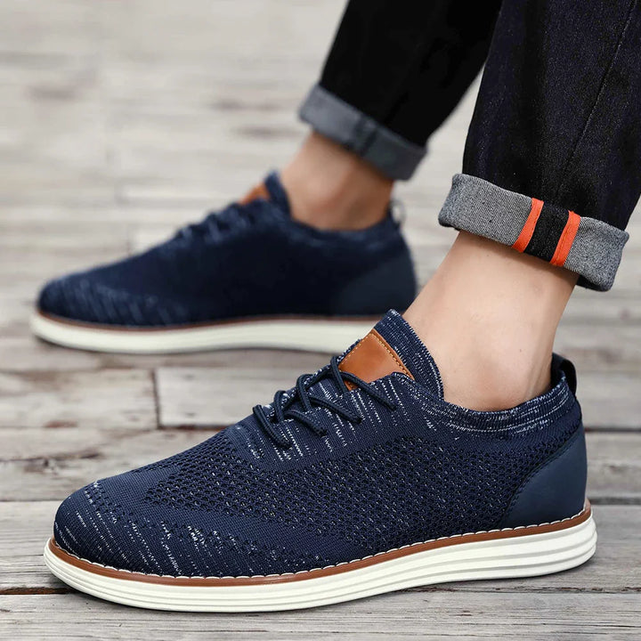 A person in casual style sports dark blue breathable knit sneakers with white soles and orange accents on a wooden floor. Their jeans are cuffed, revealing orange and gray striped socks.
