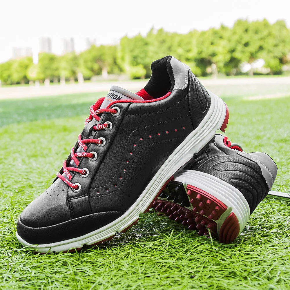 A pair of black athletic golf footwear with white soles and red accents is displayed on green grass, with trees and buildings blurred in the background. One shoe stands upright while the other is tilted, highlighting its advanced cleat technology.