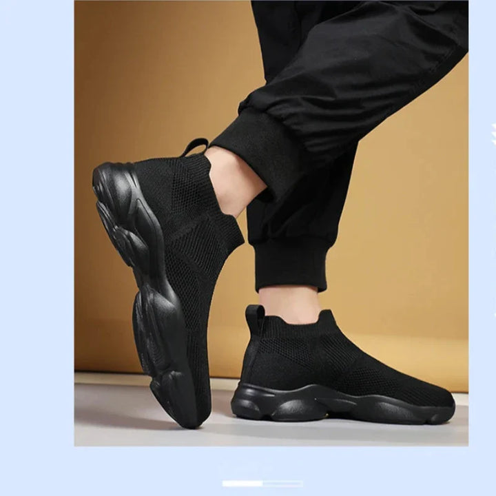 A person wearing black slip-on athletic sneakers with a non-slip sole and black jogger pants stands on a light-colored floor against a beige background. The shoes feature a chunky sole and a breathable knit fabric upper.