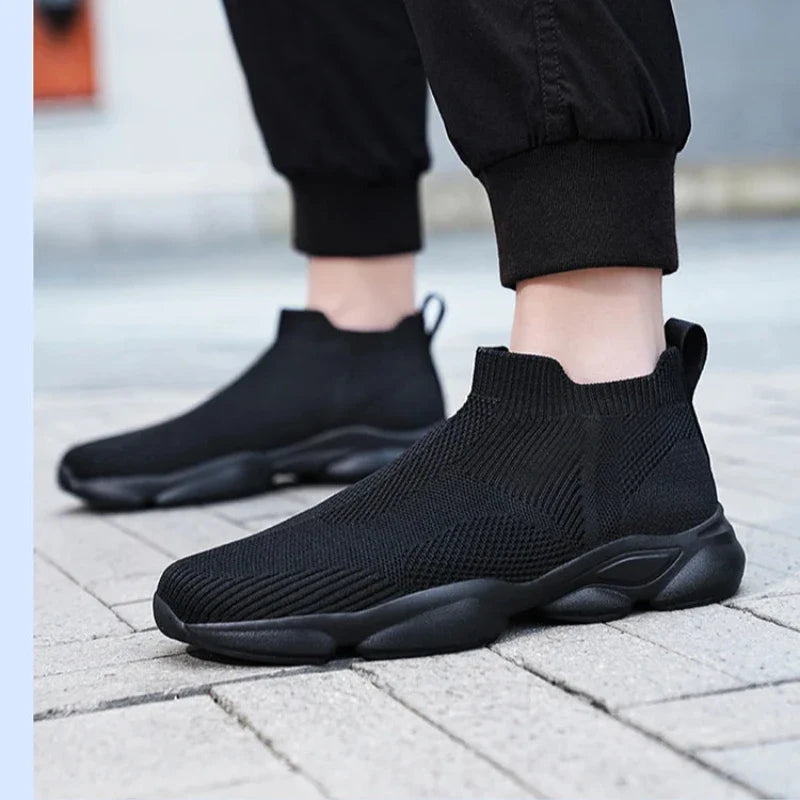 A person wears black slip-on sneakers made of lightweight, breathable knit fabric with thick non-slip soles, standing on a light-colored paved walkway. They’re dressed in black pants with elastic cuffs.