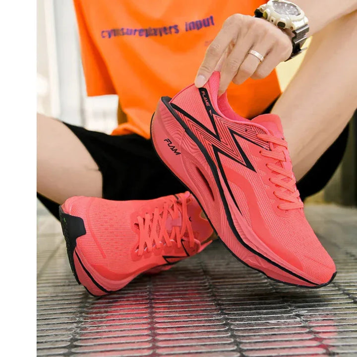 A person wearing black shorts and an orange t-shirt sits on a metal grid floor, holding up one bright pink running shoe with a black zigzag design and shock-absorbing sole, while the other ultra-lightweight shoe is worn on their foot.