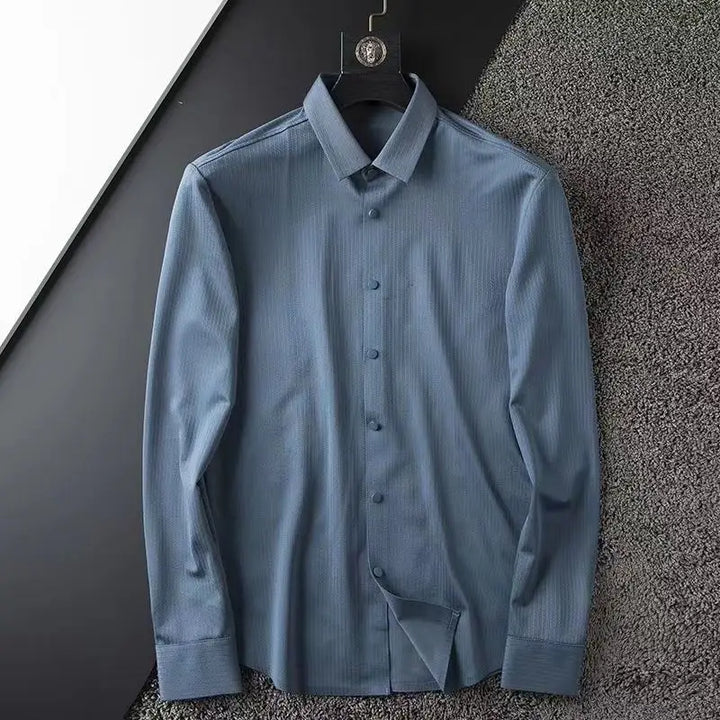 A business casual shirt, this light blue, long-sleeve dress shirt with subtle texture hangs on a black hanger against a textured grey surface. It features a classic collar and button-down front—perfect for spring.