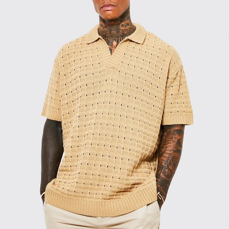 A person wears a beige short-sleeve summer shirt with a textured, perforated pattern and breathable knit design. Tattoos cover their neck and arms, complemented by light beige pants. The background is plain and light gray.