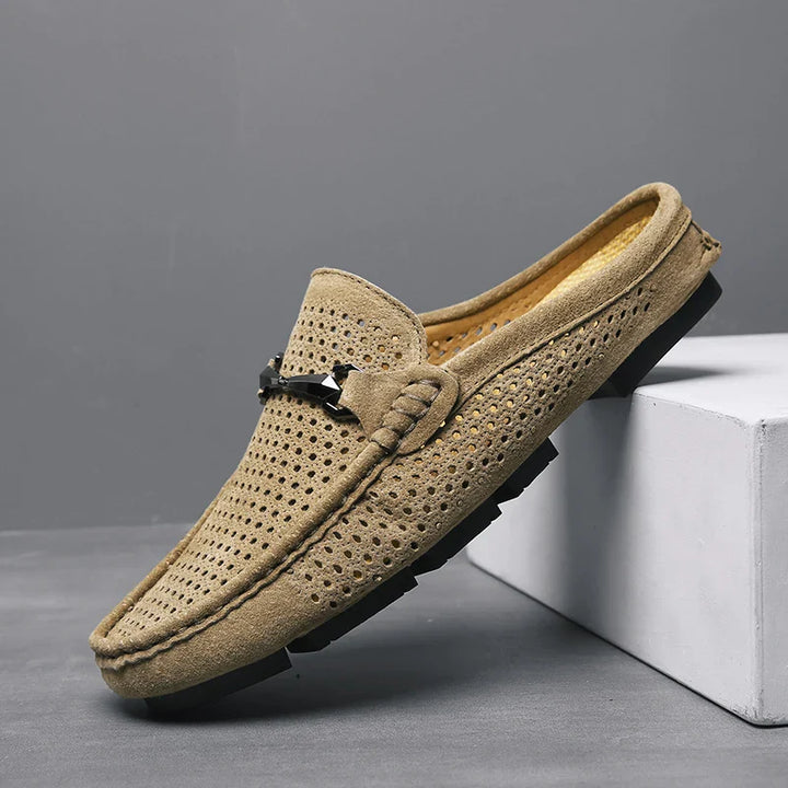 A single tan suede loafer with perforated detailing, innovative cushioning, black rubber sole, and a metal buckle accent, displayed against a gray background and propped on a white block.