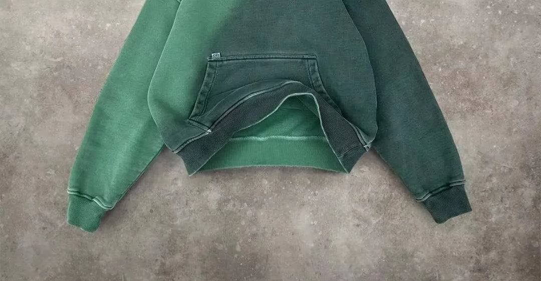 A green, relaxed fit sweatshirt with a front pocket is laid flat on a textured gray concrete surface. The breathable fabric appears slightly worn, with visible seams and faded edges.