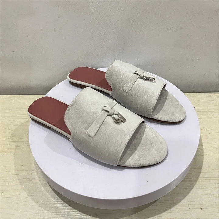 A pair of light beige, open-toe slip-on sandals with flat soles, a cushioned insole, and a decorative bow with metal accent on top, displayed on a round white platform against a plain background.