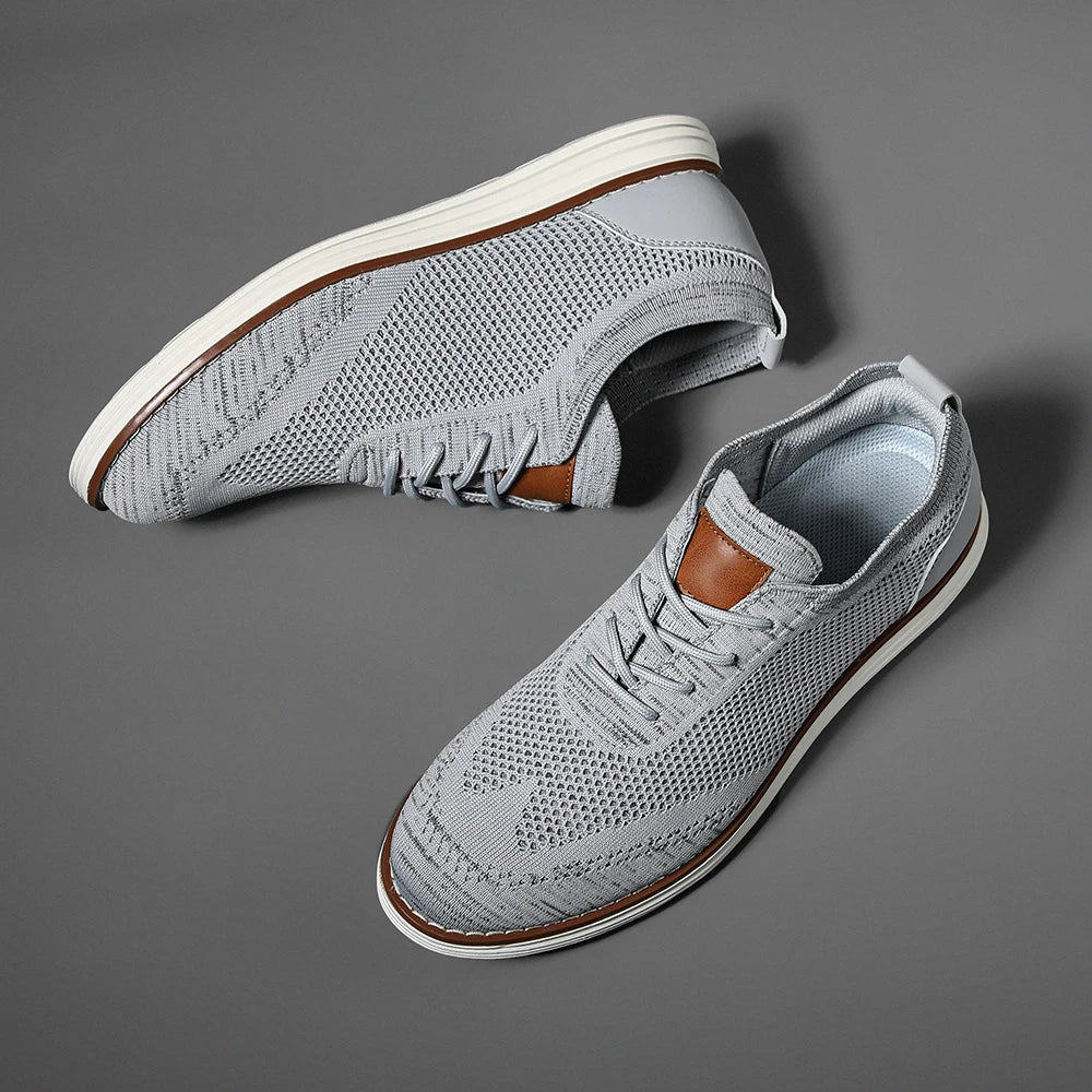 A pair of light gray breathable knit sneakers with white soles and brown accents on the tongue and heel, displayed on a flat gray surface. These stylish shoes feature a modern, sporty design perfect for casual style.