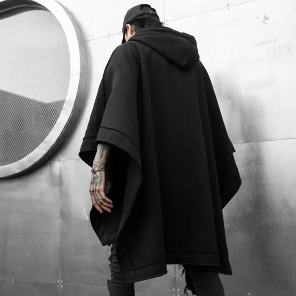A person in a loose, black Techwear hoodie stands with their back to the camera against a metallic wall featuring a large circular mesh detail. Tattoos are visible on one hand, and they wear a black cap, embracing streetwear vibes.