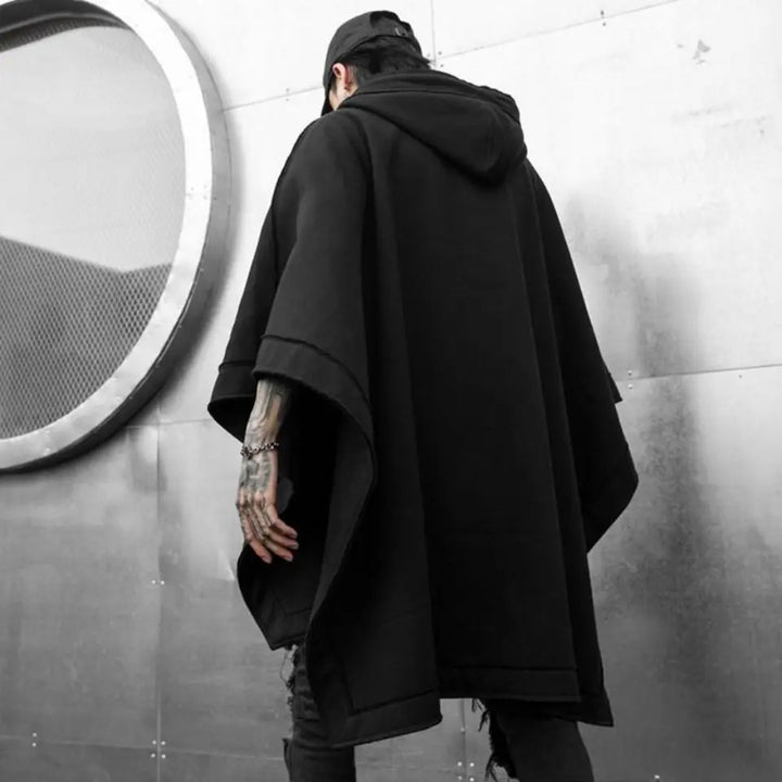 A person in a loose, black Techwear hoodie stands with their back to the camera against a metallic wall featuring a large circular mesh detail. Tattoos are visible on one hand, and they wear a black cap, embracing streetwear vibes.