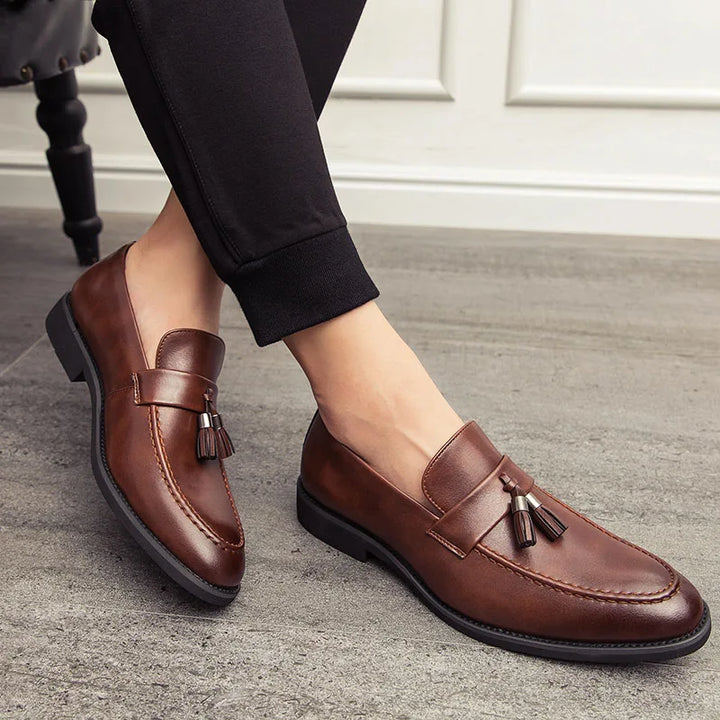 A person wearing polished leather tassel loafers sits indoors on a gray floor. The brown loafers with black soles complement the minimalistic shoes style, paired perfectly with classic black pants.