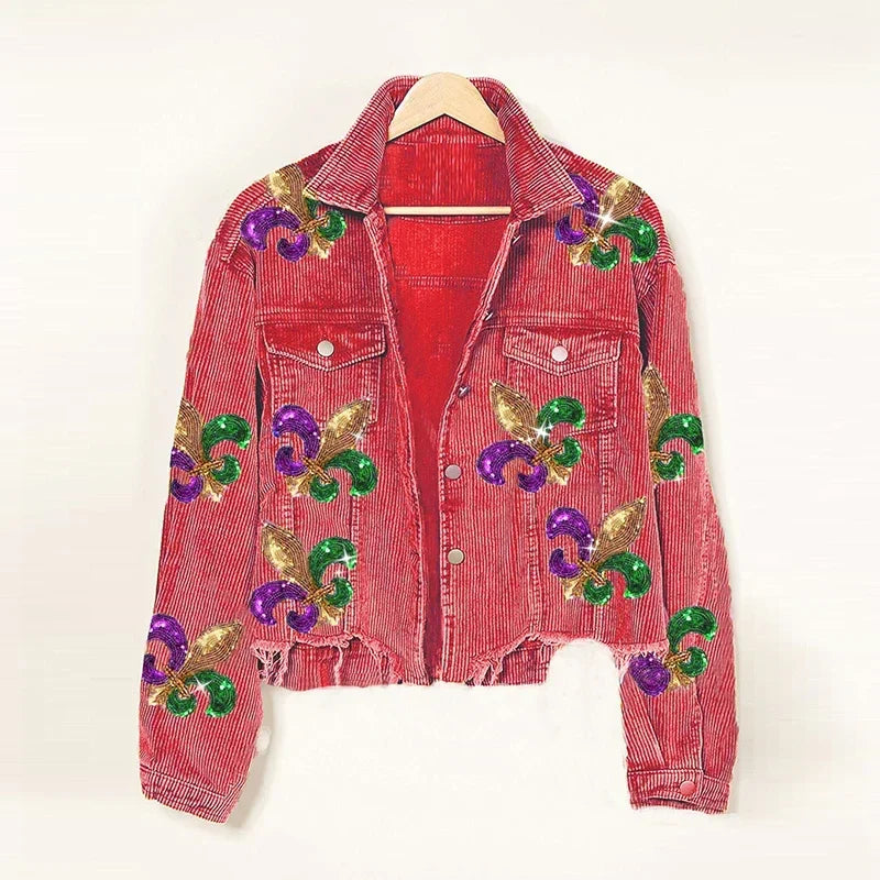 A red corduroy jacket hangs on a wooden hanger, featuring a cropped relaxed fit and large sequin fleur-de-lis designs in green, purple, and gold. Detailed with buttons, chest pockets, and a slightly frayed hem.