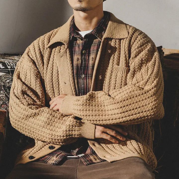 A man sits with his arms crossed, wearing a chunky knit beige cardigan over a plaid shirt and brown pants. His face is partially out of frame, and the lighting creates a warm, cozy heritage aesthetic.