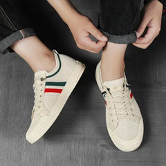 A person wearing beige sneakers with a breathable mesh upper and red and green stripes is rolling up the cuff of their dark jeans while sitting on a dark floor.