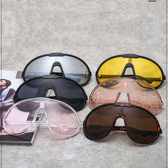 Five pairs of oversized sunglasses in bold colors—black, yellow, clear, pink, and tortoiseshell brown—are arranged on a grey surface next to a partially visible open magazine, perfect for outdoor activities and featuring UV400 protection.