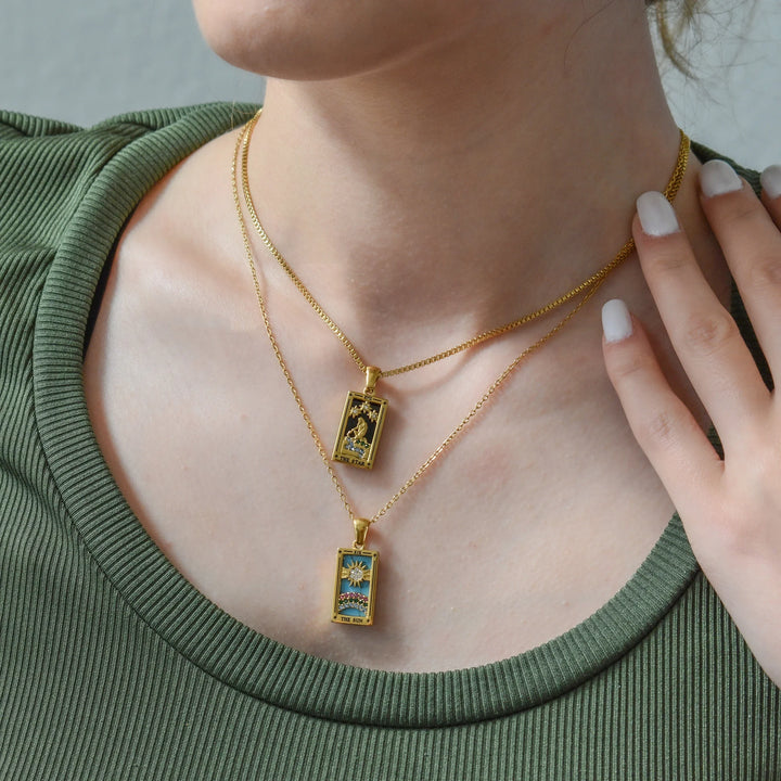 A person wearing an olive green ribbed shirt models two layered, 18K gold-plated Solaris Pendant Necklaces with colorful, rectangular designs featuring Blue Enamel; their hand rests near their neck, showing white polished nails.