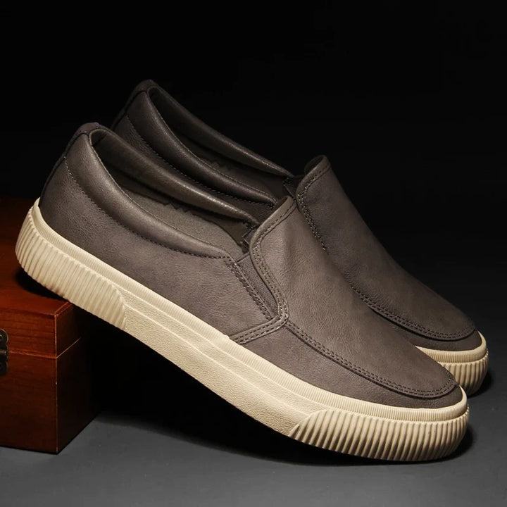 A pair of men’s slip-on casual shoes with dark gray uppers and thick, textured hybrid rubber soles, displayed against a dark background with one shoe propped on a wooden box.
