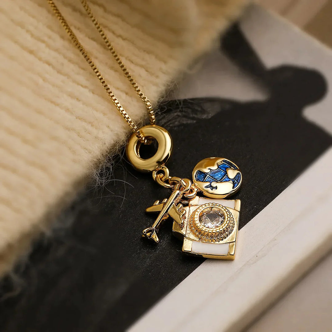 A gold adventure necklace with a delicate chain features three charms—a blue and white airplane, a rhinestone-adorned vintage camera, and a small key—resting on beige knitted fabric and a black-and-white surface.