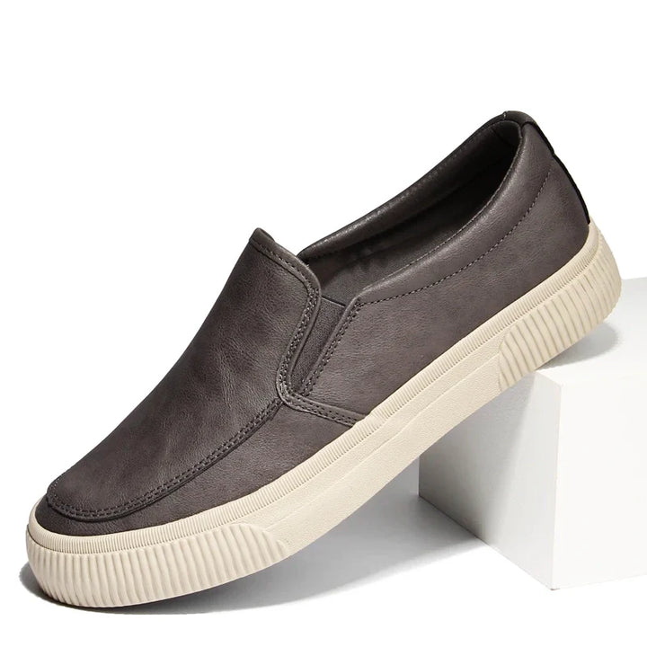A single gray men’s slip-on sneaker with a hybrid rubber sole and white textured finish is displayed, propped at an angle on a white block against a plain background.