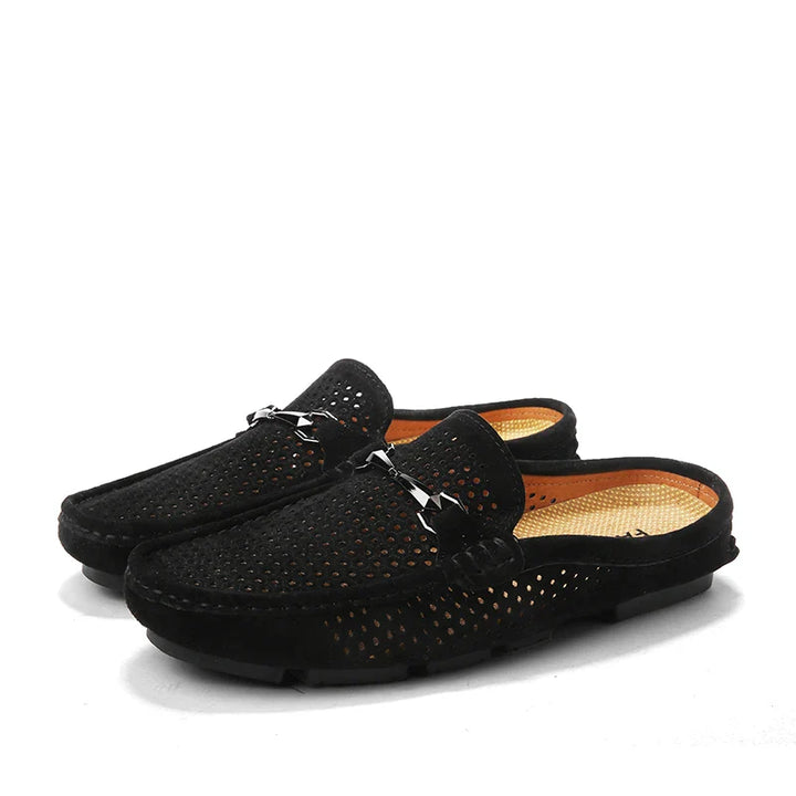 A pair of black slip-on breathable loafers with perforated uppers, open backs, and metal buckle details on the fronts. The shoes have tan insoles and black soles, making them perfect summer shoes.