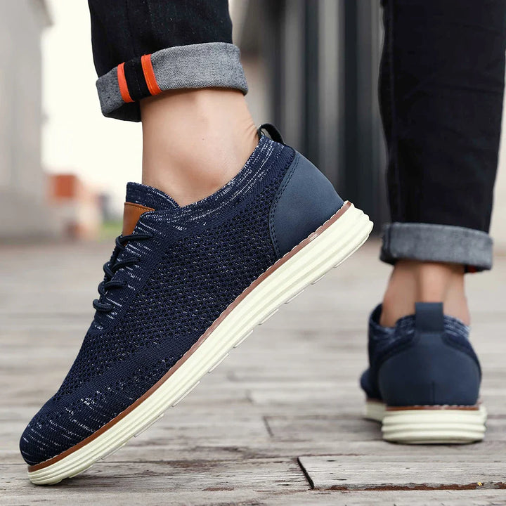A person wearing dark blue breathable knit sneakers with white soles stands on a wooden surface outdoors. The person’s jeans are rolled up, revealing the shoes and part of their ankles for a relaxed casual style.