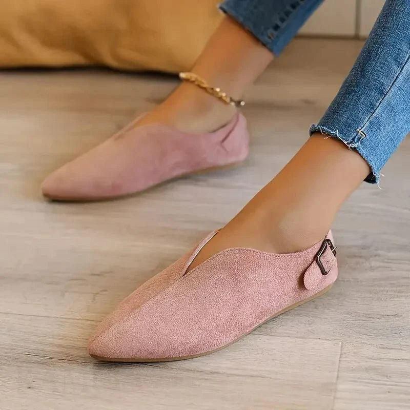 A person wearing pink suede pointed toe shoes with a small buckle detail and cushioned insole sits on a light wooden floor, dressed in blue skinny jeans and an anklet.