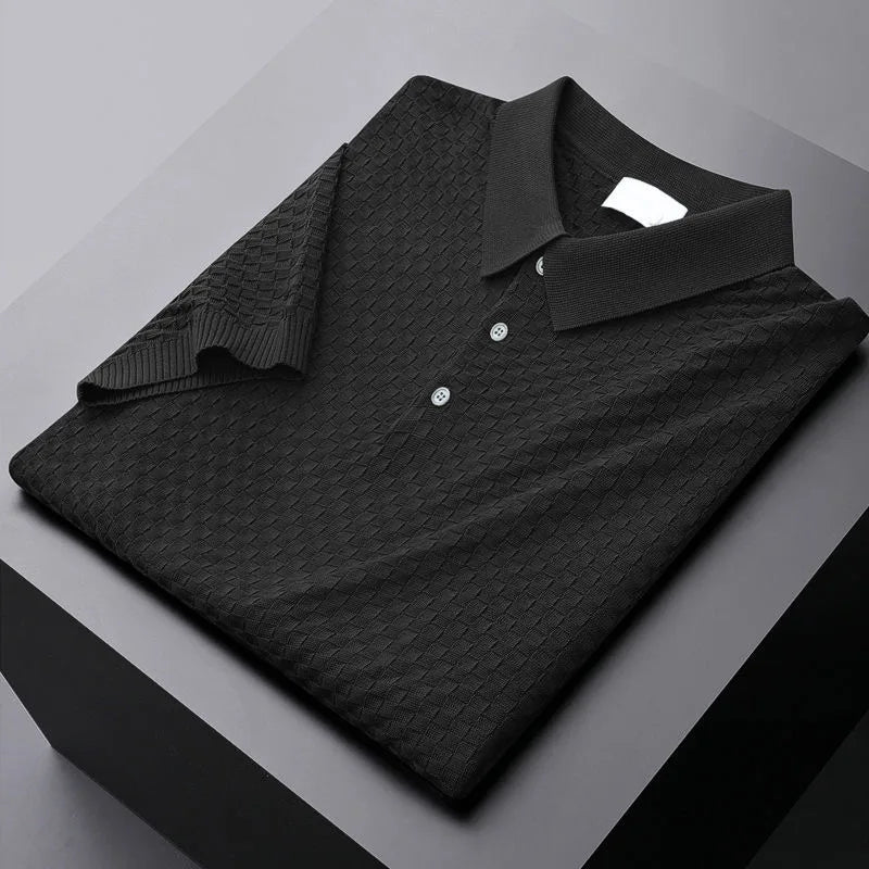A neatly folded black polo shirt with a textured waffle jacquard diamond pattern, three buttons, and a classic collar, placed on a sleek, gray, geometric surface.