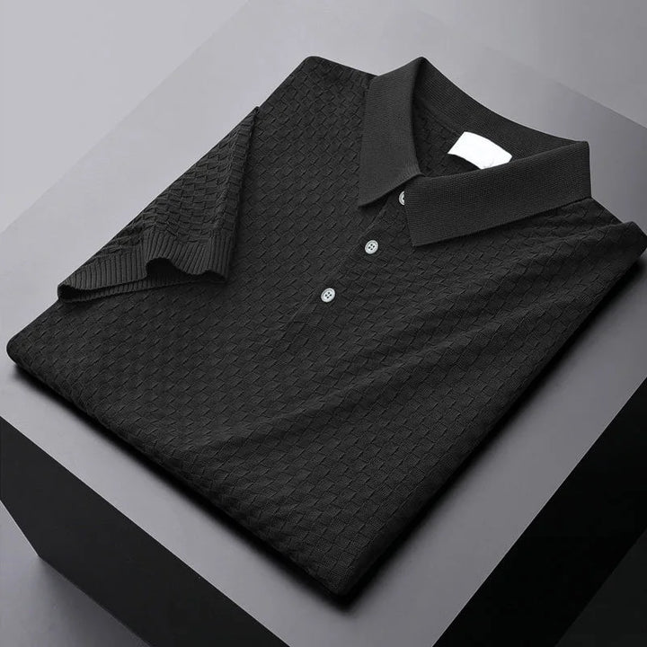 A neatly folded black polo shirt with a textured waffle jacquard diamond pattern, three buttons, and a classic collar, placed on a sleek, gray, geometric surface.