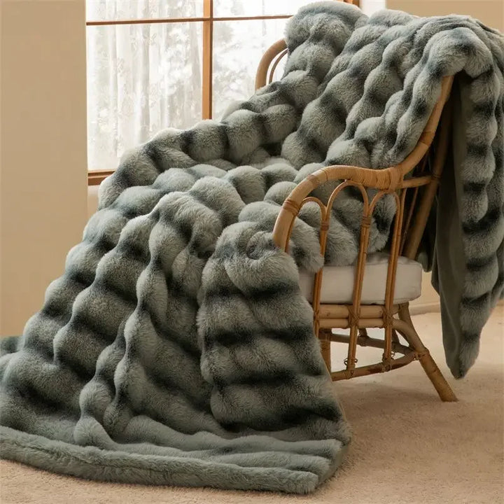 A soft, gray faux rabbit fur blanket with an elegant wave pattern draped over a cushioned rattan chair next to a window with sheer curtains creates a cozy and inviting atmosphere.
