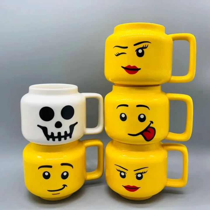 Five stackable coffee mugs are arranged in a pyramid; four are yellow with different smiling cartoon faces, and one is white with a black skull face. These fun, Lego-inspired mugs stand out against a plain gray background.