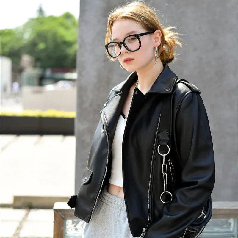 A young woman with light hair wearing large black glasses, a black leather jacket with zipper pockets, white crop top, and gray pants stands outdoors, looking at the camera with a neutral expression. Trees and buildings are blurred in the background.