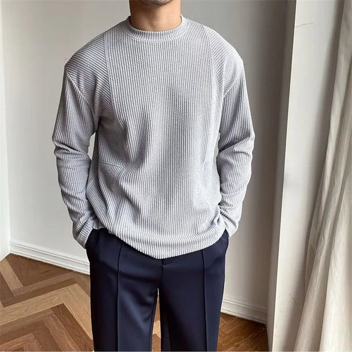 A person stands indoors in a light grey, ribbed knit Grayson Crew Classic sweater and dark pants, hands in pockets. The background features a white wall and a window with a light curtain.