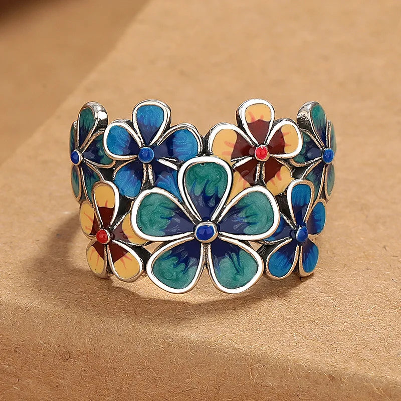 A wide silver ring with vibrant colors, featuring enamel flowers in blue, yellow, and green—each with a red or blue center—displayed on a light brown surface.