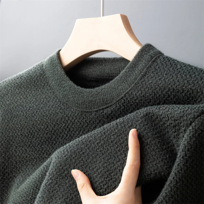 A hand holding a dark green high-quality cotton sweater on a wooden hanger, shown close-up against a plain gray background.