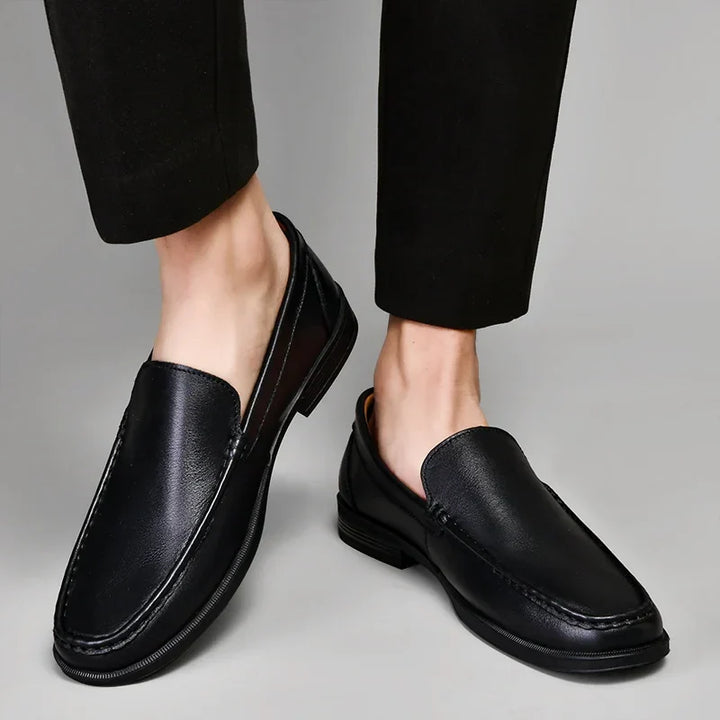 A person wearing black dress pants and classic slip-on loafers made from genuine leather stands on a light gray surface, with one foot slightly raised, showcasing the sleek design of these timeless shoes.