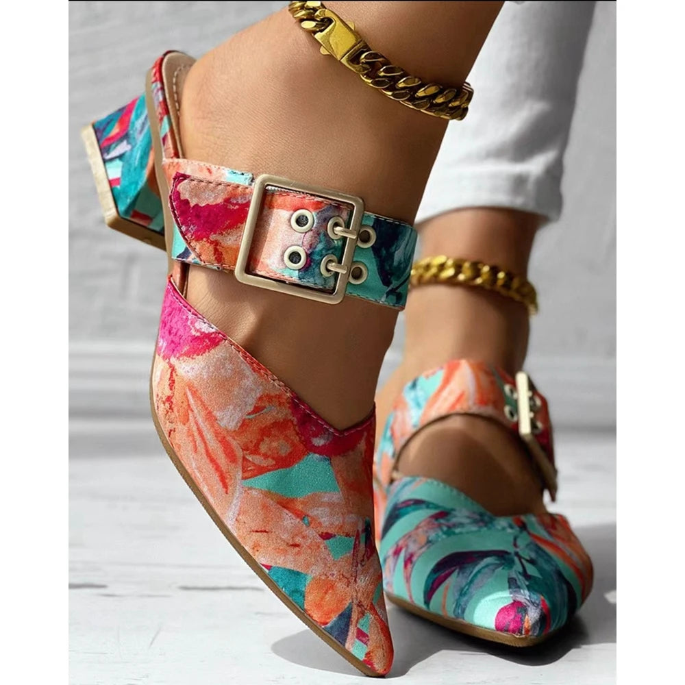 A close-up of feet in colorful, floral-patterned square heel slippers with large buckles and chunky heels. Paired with white pants and a gold chain anklet, these summer mules feature vibrant pink, teal, orange, and cream tones.