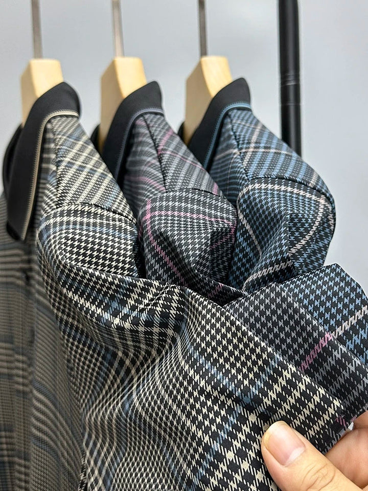 Three plaid patterned shirts in shades of gray, black, and blue hang on wooden hangers. A hand in the foreground holds and slightly bunches the breathable fabric of the blue-accented shirt.