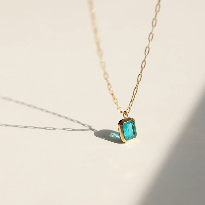 A delicate 18K gold plated chain necklace with a rectangular blue gemstone pendant rests on a light surface, casting a soft shadow.