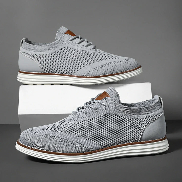 A pair of light gray, breathable knit sneakers with white soles, subtle brown accents on the tongue, and white laces, displayed against a gray background with one shoe elevated on a white block for effortless casual style.