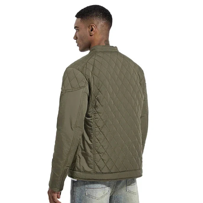 A man wearing a wind-resistant quilted olive green slim-fit jacket stands facing away, showing the back and side of the jacket. He is also wearing light blue jeans. The background is plain white.